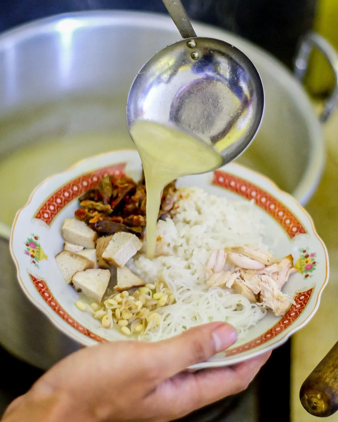 Someone pouring broth over a bowl of Nasi Lesah with a ladle